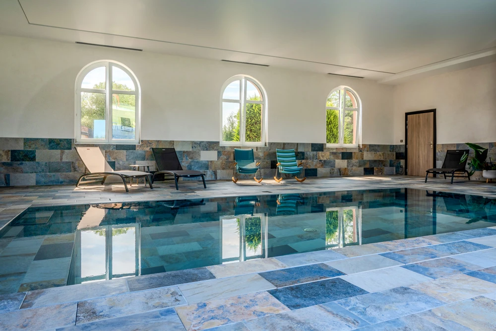 Piscine intérieure rectangulaire de type couloir de nage, carrelage en pierre naturelle multicolore aux tons bleus et ocres, murs en enduit blanc avec frise en pierre, trois fenêtres cintrées, transats noirs, chaises longues à bascule en bois